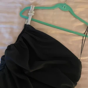 Svart axelbandslös klänning - Elegant svart axelbandslös klänning med en glittrig axelrem. Klänningen har en smickrande passform och en stilren design, perfekt för festliga tillfällen. Den har en knälång längd och en subtil slits för extra stil.