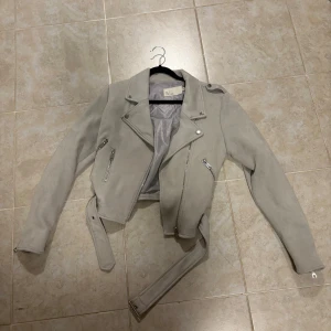 Grå mockajacka från NLY Trend - Snygg grå mockajacka från NLY Trend med dragkedjor och bälte. Perfekt för en stilren look. Jackan har långa ärmar och en klassisk bikerstil. Använd ett flertal gånger, fint skick.