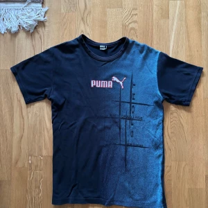 Svart t-shirt från Puma - Snygg svart t-shirt från Puma med tryck i rött och svart. Perfekt för sportiga aktiviteter eller en avslappnad stil. Tillverkad i ett bekvämt material med korta ärmar och rund hals.