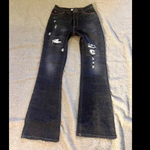 Svarta jeans med slitningar - Snygga svarta lågmidjade  jeans med slitningar på framsidan och coola sömmar på bakfickorna. De har en bootcut-stil och är perfekta för en avslappnad look. Jeansen har röda sömmar som ger en extra touch.