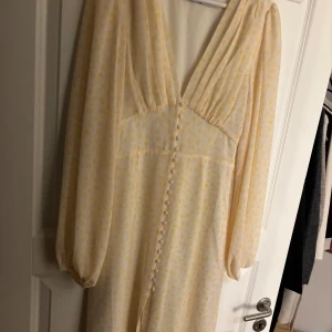 Gul blommig klänning med knappar - Charmig gul klänning med blommigt mönster och knappar framtill. Den har långa ärmar och en v-ringad design som ger en romantisk känsla. Perfekt för vår och sommar!