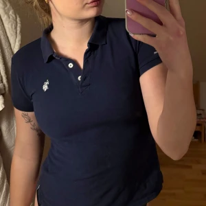 Mörkblå pikétröja från U.S. Polo Assn. - Säljer en stilren mörkblå pikétröja från U.S. Polo Assn. med klassisk krage och knappar framtill. Tröjan har en liten broderad logotyp på bröstet och är tillverkad i 100% bomull för en bekväm passform.