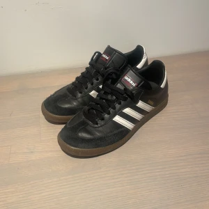 Svarta Adidas Samba sneakers - Snygga svarta Adidas Samba sneakers med vita ränder och brun sula. Skorna har en klassisk design med snörning och är tillverkade i skinn och mocka. Perfekta för en stilren look. Mockan är något sliten men inget som stör