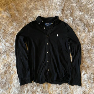 Ralph Lauren featherweight mesh - Snygg svart långärmad skjorta från Ralph Lauren featherweight mesh med klassisk krage och knappar framtill. Den har en broderad logga på bröstet som ger en stilren touch. Perfekt för en stilmedveten look. Passar till alla outfits och lager på lager outfits!