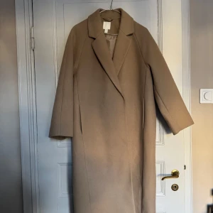 Beige kappa - Säljer en stilren beige kappa med klassisk krage och långa ärmar. Perfekt för höst och vinter. Kappan har en enkel design. Lite nopprig men inget som förstör 