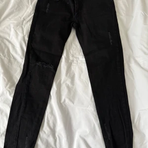 Svarta jeans med slitningar från House of Lola - Snygga svarta jeans med slitningar på låret och knäna. Oanvända men saknar prislapp. De har en tight passform med mycket stretch och ör lite lägre i midjan. Jeansen har dragkedja och knapp framtill samt bakfickor. Byxorna räcker till anklarna.