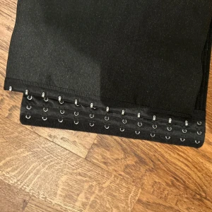 Svart corsett - Snyggt svart corsett med hyskor för justering. Perfekt för att ge en stilren look till din outfit. Bältet är brett och ger bra stöd.