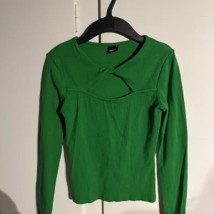 Grön ribbad topp från Gina Tricot - Säljer en snygg grön ribbad topp från Gina Tricot med unik detalj vid halsen. Toppen har långa ärmar och är perfekt för en stilren look. Passar bra till både jeans och kjol. 🌿