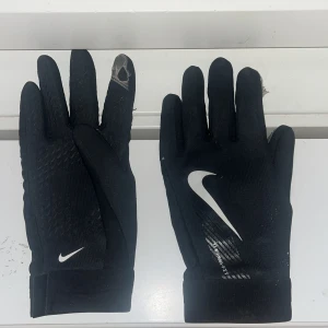Svarta handskar från Nike - Säljer ett par svarta handskar från Nike med deras ikoniska logotyp i vitt. Handskarna har en texturerad yta för bättre grepp och är designade för att hålla händerna varma under kalla dagar. Perfekta för vinteraktiviteter eller löpning i kyligt väder. ( slitna på pekfingret och tummen)