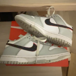 Nike Dunk i vitt och grått - Snygga Nike Dunk sneakers i vitt och grått med svart swoosh. Skorna har en klassisk design med snörning och perforerade detaljer på tån för extra ventilation. Perfekta för en stilren look. +skrapbar logga, alltså att man kan skrapa nike loggan till röd färg