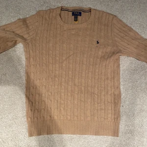 Beige stickad tröja från Ralph Lauren - Säljer en snygg beige stickad tröja från Ralph Lauren med kabelstickat mönster. Tröjan har långa ärmar och en broderad logga på bröstet. Perfekt för en stilren look. Använt ändast en gång, pris går såklart att diskutera. 