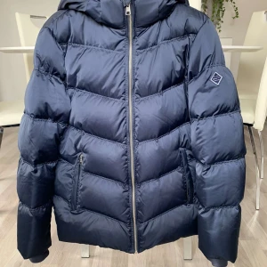 Mörkblå pufferjacka med huva - Säljer en snygg mörkblå pufferjacka med huva och dragkedja framtill. Jackan har långa ärmar och två praktiska fickor med dragkedja. Perfekt för kyliga dagar!