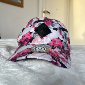Blommig keps från Upfront Company - Snygg keps från Upfront med ett blommigt mönster i rosa, vitt och svart. Kepsen har en justerbar baksida för perfekt passform och en framträdande logotyp på framsidan. Perfekt för att ge din outfit en färgglad touch!