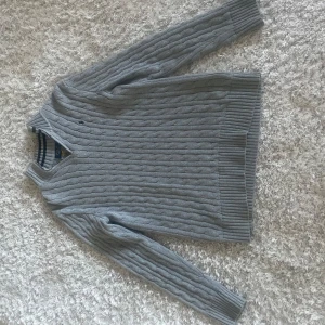Grå stickad tröja från Ralph Lauren - Säljer en stilren grå stickad tröja från Ralph Lauren med kabelstickat mönster. Tröjan har en dragkedja vid halsen och långa ärmar. Perfekt för kyligare dagar och ger en klassisk look.