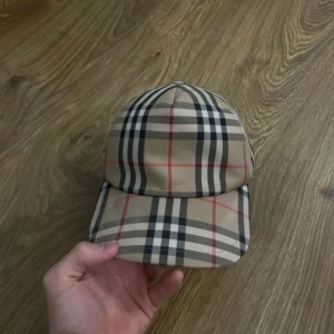 Burberry keps - Snygg rutig keps från Burberry i klassiskt beige, svart och rött mönster. Kepsen har en justerbar rem baktill och en broderad Burberry-logga. Perfekt för att ge din outfit en stilren touch.