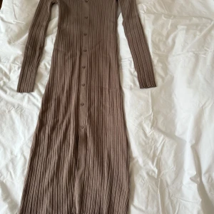  Ribbad klänning från Neo Noir i färgen Taupe - Säljer en snygg Taupe färgad långklänning från Neo Noir. Klänningen är ribbad med långa ärmar och knappar framtill. Perfekt för en stilren look. Tillverkad i en mjuk blandning av viskos och nylon.
