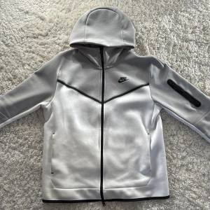 Grå/vit hoodie från Nike - Säljer en stilren grå/vit hoodie från Nike med dragkedja och huva. Hoodien har en svart Nike-logga på bröstet och en praktisk ficka med dragkedja på ärmen. 