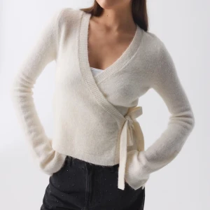 Vit omlottkofta från Gina - Säljer en stilren vit omlottkofta från Gina. Den har en knytning i sidan och är tillverkad i ett mjukt material. Perfekt för lager-på-lager och ger en elegant touch till din outfit.