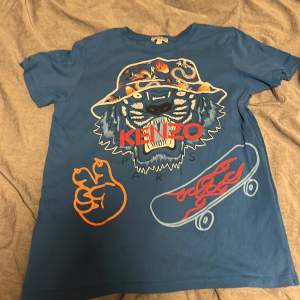 Säljer en cool blå t-shirt från Kenzo Kids. Den har ett stort tryck med en tiger som bär en hatt och en skateboard med eldflammor. Perfekt för en avslappnad stil!