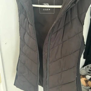 Svart quiltad dunväst från Zara - Säljer en svart quiltad dunväst från Zara. Västen har en dragkedja framtill och är perfekt för kyligare dagar. Den är stilren och passar till det mesta i garderoben. Perfekt för höst och vinter!