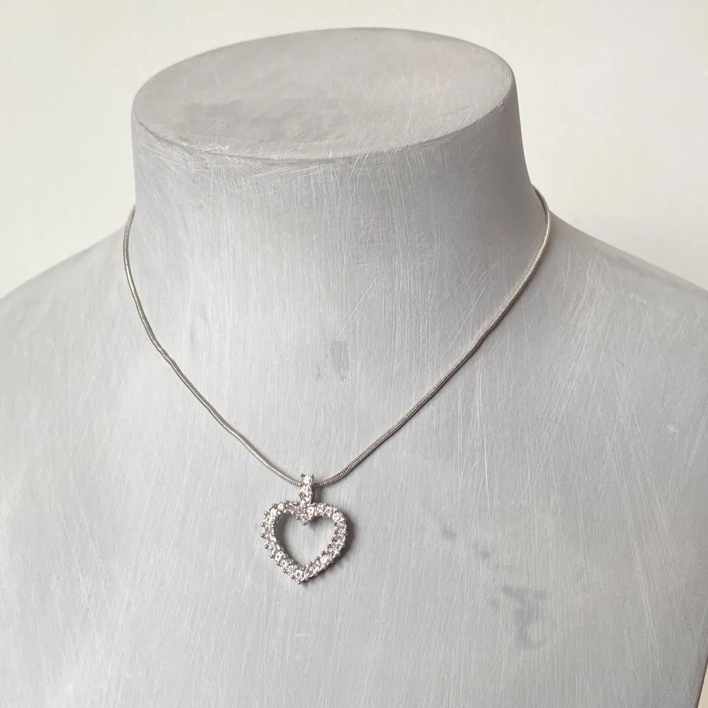 Säljer ett elegant silverhalsband med en glittrig hjärtformad berlock. Halsbandet har en tunn kedja och berlocken är täckt med små gnistrande stenar som ger en lyxig känsla. Perfekt för både vardag och fest! 💖. Accessoarer.