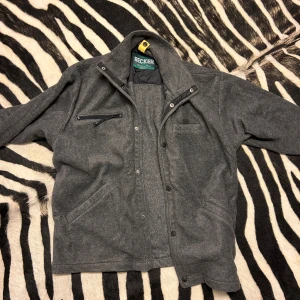Grå jacka från Recker - Säljer en snygg grå fleece jacka från Recker i storlek medium. Jackan har en dragkedja och knappar framtill, vilket ger den en stilren look. Perfekt för höst och vår med sitt mjuka material och praktiska fickor. 