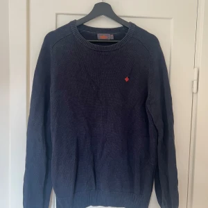 Mörkblå stickad tröja från Morris - Säljer en snygg mörkblå stickad tröja från Morris. Den har en liten röd broderad detalj på bröstet och ribbstickade muddar vid ärmslut och nederkant. Perfekt för kyliga dagar!