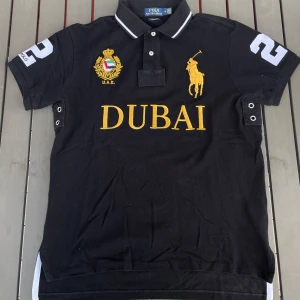 Svart polo från Ralph Lauren med Dubai-tryck - Säljer en snygg svart polo från Ralph Lauren med stort gult Dubai-tryck på framsidan. Tröjan har korta ärmar och en klassisk krage med vita detaljer. På bröstet finns ett broderat emblem med U.A.E. och en stor gul polospelare. Siffran 2 är tryckt på både ärmen och ryggen. Perfekt för en stilren och sportig look!