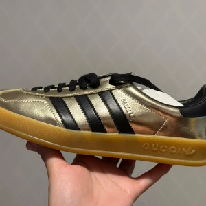 Guldiga Adidas Gazelle x Gucci sneakers oanvända - Unika Adidas Gazelle sneakers i samarbete med Gucci. Skorna har en glittrig guldfärgad ovandel med svarta ränder och en gummisula med Gucci-logotyp. Perfekta för den som vill sticka ut med stil.
