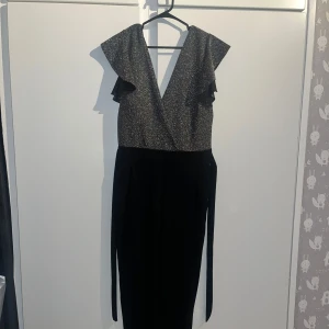 Glittrig svart byxdress med volangdetaljer - Elegant svart byxdress med glittrig överdel och volangdetaljer vid axlarna. Den har ett strechig material och en omlott-design och är ärmlös, vilket ger en stilren look. Perfekt för festliga tillfällen.