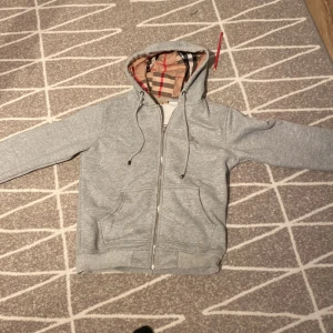 Grå hoodie från Burberry - Säljer en stilren grå hoodie från Burberry med klassiskt rutigt mönster i huvan. Tröjan har dragkedja framtill och justerbara snören i huvan. Perfekt för en avslappnad och trendig look.
