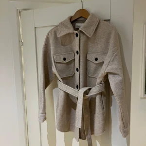Beige jacka från NLY Trend - Snygg beige overshirt/jacka från NLY Trend med knappar och bälte i midjan. Jackan har två stora bröstfickor med lock och långa ärmar. Perfekt för lager-på-lager-stil under hösten.