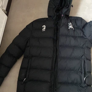 Polo jacka - Snygg svart dunjacka med broderad logga på bröstet och nummerdetalj. Jackan har en huva och dragkedja framtill. Perfekt för kyliga dagar.