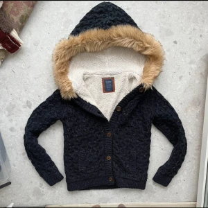 Tröja med päls från bershka - Mysig svart stickad jacka från Bershka med knappar och en stor huva kantad med fuskpäls. Perfekt för kyliga dagar med sin varma och bekväma design.