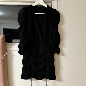 Svart klänning från Gina Tricot - Elegant svart klänning från Gina Tricot med v-ringning och puffiga ärmar. Klänningen har en smickrande omlott-design och en dragkedja i sidan för enkel påklädning. Perfekt för en stilren look.