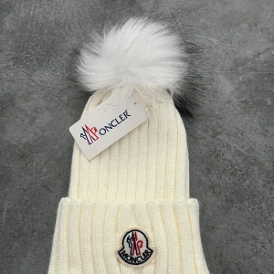 Vit stickad mössa - Snygg vit stickad mössa från Moncle med en fluffig pälsboll på toppen. Mössan har en ribbad design och Moncler-loggan framtill. Perfekt för att hålla sig varm och stilren under kalla dagar.