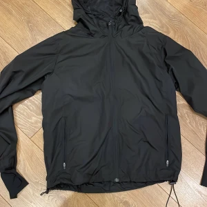 Svart vindjacka från TriDri - Säljer en svart vindjacka från TriDri i storlek M. Jackan har dragkedja framtill och justerbar huva. Perfekt för blåsiga dagar med sin lätta och funktionella design.