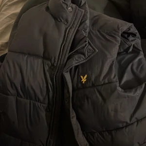 Svart dunjacka från Lyle & Scott - Snygg svart dunjacka från Lyle & Scott med dragkedja och knappar. Jackan har en klassisk design med det ikoniska guldlogotypen på bröstet. Perfekt för kyliga dagar.