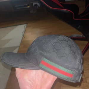 svart gucci keps med mönster och ränder - Snygg svart keps från Gucci med diskret mönster och klassiska gröna och röda ränder på sidan. Perfekt för en stilren look.