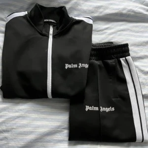 Svart träningsjacka från Palm Angels - Snygg svart träningsjacka från Palm Angels med vit dragkedja och vita ränder längs ärmarna. Perfekt för en sportig look eller avslappnad stil. Jackan har en hög krage och är tillverkad i ett bekvämt material.