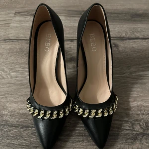 Svarta pumps med kedjedetalj från BEBO - Eleganta svarta pumps från BEBO med spetsig tå och klack. Skorna har en iögonfallande kedjedetalj i silver runt öppningen, vilket ger en lyxig touch. Perfekta för att lyfta vilken outfit som helst.