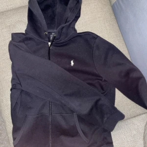 Svart hoodie från Ralph Lauren - Snygg svart hoodie från Ralph Lauren med dragkedja och klassisk logga på bröstet. Köpt från Zalando men använd max 2-3 gånger. Passar L/XL