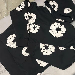 Svart hoodie med vita blommor från Denim Tears - Säljer en svart hoodie från Denim Tears med vita blommönster. Den har en klassisk passform med huva och långa ärmar. Perfekt för en avslappnad stil.