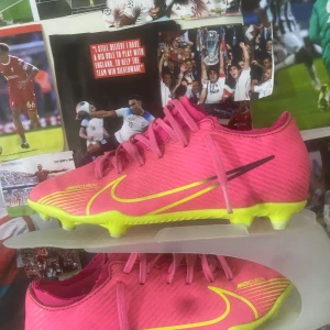 Rosa Nike Mercurial fotbollsskor - Säljer ett par rosa Nike Mercurial fotbollsskor med gula detaljer och snörning. Skorna har en neonfärgad sula med dobbar, perfekt för gräsplaner. De är designade för snabbhet och kontroll på planen. Säljer dessa för att jag rensar ut min fotbolls kollektion 