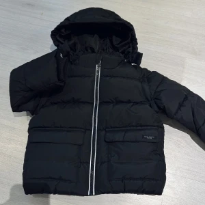 Svart pufferjacka med avtagbar huva - Säljer en svart pufferjacka med avtagbar huva och praktisk dragkedja framtill. Jackan har långa ärmar och stora fickor för extra förvaring. Perfekt för kyliga dagar!Helt ny jackan för barn . Passar perfekt för 24-36 månader