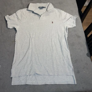 Grå pikétröja från Ralph Lauren - Säljer en stilren grå pikétröja från Ralph Lauren med klassisk krage och korta ärmar. Tröjan har en broderad logotyp i vinrött på bröstet. Perfekt för en avslappnad men ändå snygg look.