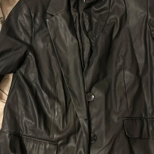 Svart skinnjacka från NA-KD - Snygg svart skinnjacka kavaj från NA-KD med klassisk design och knappar framtill. Perfekt för en stilren look. Jackan har långa ärmar och två fickor framtill. Använd en gång