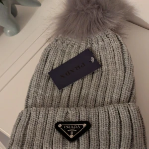 Grå ribbad mössa från Prada - Snygg grå ribbad mössa från Prada med en fluffig pälsboll på toppen. Perfekt för att hålla dig varm och stilren under kalla dagar.