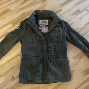 Grön field jacket! - Snygg grön jacka från Khujo med flera fickor och coola patchar på ärmarna. Jackan har dragkedja och knappar framtill samt en hög krage. Jackan har mycket snygga och unika detaljer och är i mycket bra skick. Kan passa S