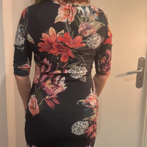 Blommig klänning från Sisters Point - Snygg blommig klänning från Sisters Point med trekvartsärmar och en figurnära passform. Klänningen har ett färgglatt mönster med stora blommor i rött, rosa och vitt på en svart bakgrund. Perfekt för en färgglad look!